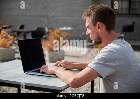 Primo piano di un giovane bell'uomo che lavora o studia in un computer portatile all'aria fresca nel bar. Concetto di processo che lavora nel computer. Foto Stock