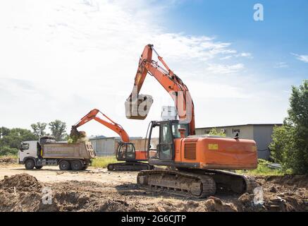 due escavatori cingolati arancioni e un dumper grigio in fase di scavo. Preparazione del sito. Carico e trasporto di suolo Foto Stock