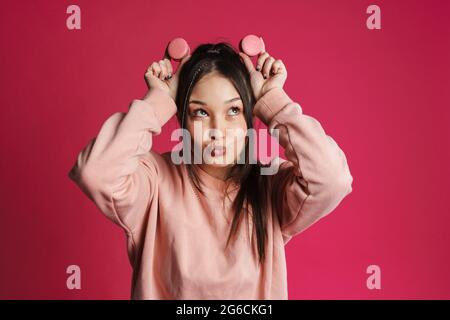 Asian brunette donna felice che si diverte con i macaroni isolati sopra la parete viola Foto Stock