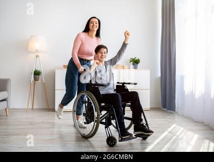 Cool mamma giovane e suo figlio disabile in sedia a rotelle divertirsi a casa, a tutta lunghezza. Concetto di passatempi figlio padre Foto Stock