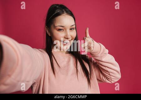 Donna asiatica felice che punta il dito verso la fotocamera mentre scatta foto selfie isolato sopra la parete viola Foto Stock