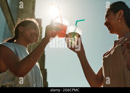 Ritratto ad angolo basso di due giovani donne che in estate si aggraffano a calici da cocktail contro il cielo azzurro illuminato dal sole Foto Stock