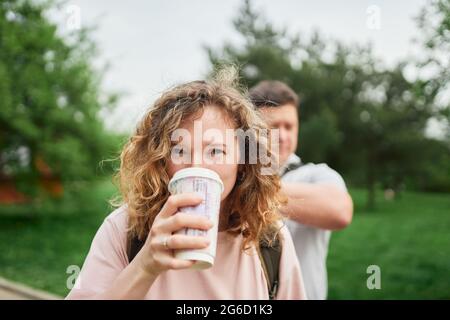 Affascinante donna con capelli ricci gustando una bevanda calda in tazza di carta da andare mentre si guarda la macchina fotografica nel parco estivo Foto Stock