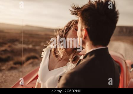 Felici sposi e sposi che si abbracciano e si baciano mentre si appoggiano su un veicolo di lusso rosso durante il viaggio su strada attraverso il Parco Naturale di Bardenas Reales a Na Foto Stock