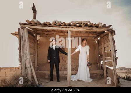 Uomo e donna del corpo completo in abiti eleganti che tengono le mani mentre si trova in piedi in un riparo di legno intemperiato il giorno del matrimonio nel Parco Naturale di Bardenas Reales in Foto Stock