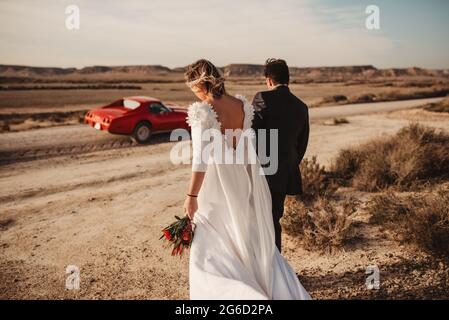 Vista posteriore di sposa anonima e sposo che passeggiano insieme verso auto rossa di lusso nel deserto del Parco Naturale di Bardenas Reales a Navarra, Spagna Foto Stock