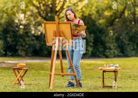 Donna pittura su tela in un parco con due tavoli laterali con attrezzi Foto Stock