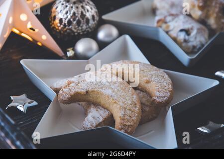 Biscotti a mezzaluna aromatizzati alla vaniglia su un piatto bianco stellato su un vassoio di legno scuro, decorato con palle di natale, stelle d'argento e stelle Foto Stock