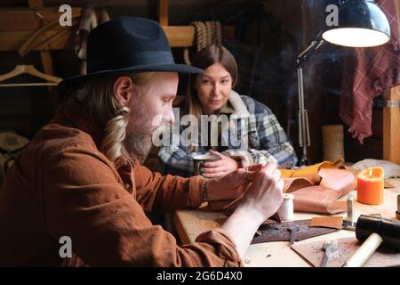 Lavoratore maturo seduto al tavolo e riparando i prodotti dalla pelle con donna seduta in background e guardando per il suo lavoro Foto Stock