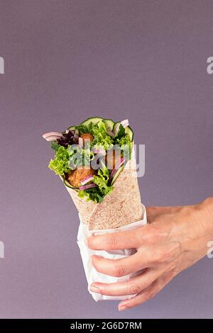 Le mani di una persona non riconoscibile che tiene un involucro di falafel vegano su sfondo viola colorato Foto Stock