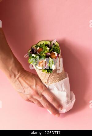 Mani di una persona non riconoscibile dal taglio corto che tiene un involucro di falafel vegano su sfondo rosa colorato Foto Stock