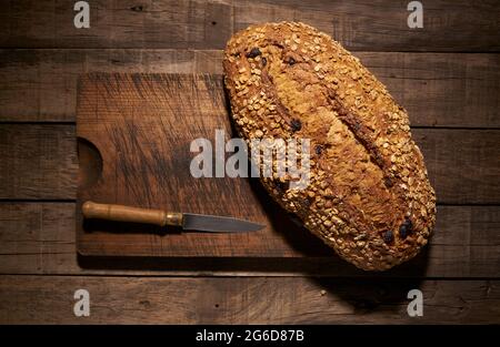 Dall'alto di un appetitoso pane fatto in casa appena sfornato con fiocchi d'avena e uvetta messo con coltello su tagliere di legno Foto Stock