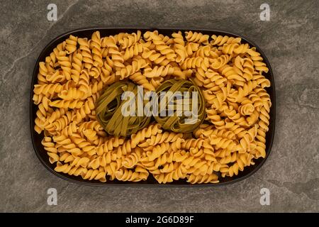 Vista dall'alto del vassoio riempito con fusilli crudi e tagliatelle di pasta e posto su un tavolo grigio Foto Stock