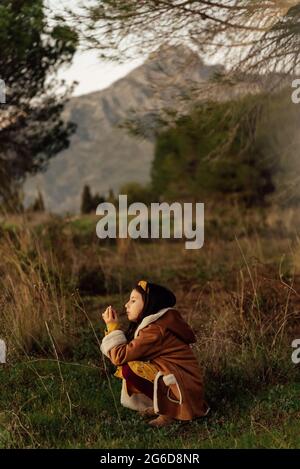 Vista laterale della bambina curiosa in abiti caldi osservando l'ambiente mentre si passa il tempo da sola sul prato erboso contro le montagne Foto Stock