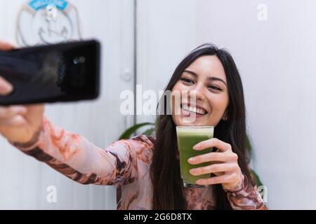 Una donna che si siede al tavolo con frullato mentre prende selfie con il telefono cellulare mentre si rilassa al bar durante il fine settimana Foto Stock