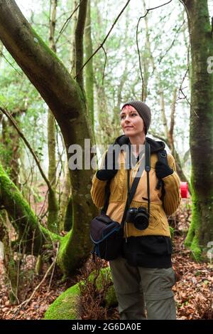 Felicissima donna in viaggio con zaino e fotocamera fotografica in piedi nei boschi verdi e guardando lontano Foto Stock