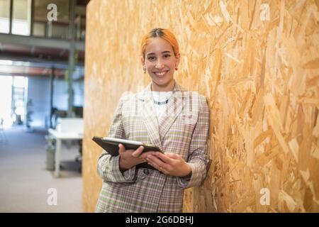 Sorridente imprenditore femminile in piedi con il computer vicino alla parete nello spazio di coworking mentre si guarda la macchina fotografica Foto Stock