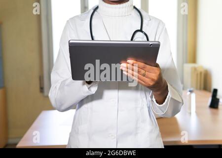 Crop giovane femmina nera medico in cappotto medico con stetoscopio che lavora con la tavoletta in un moderno studio clinico Foto Stock