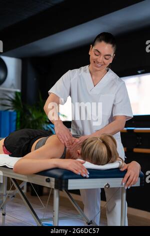 Massaggiatrice amichevole sorridendo e massaggiando le spalle della donna mentre lavora in clinica di fisioterapia Foto Stock