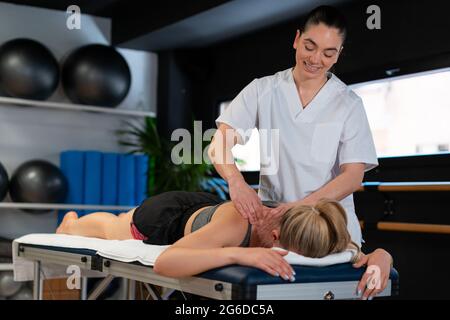 Massaggiatrice amichevole sorridendo e massaggiando le spalle della donna mentre lavora in clinica di fisioterapia Foto Stock