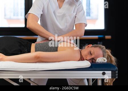 Massaggiatrice tagliata irriconoscibile sorridendo e massaggiando le spalle della donna mentre lavora in clinica di fisioterapia Foto Stock