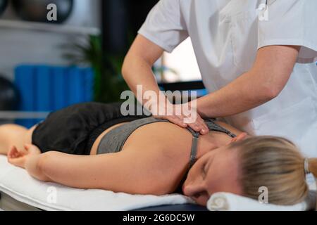 Massaggiatrice tagliata irriconoscibile sorridendo e massaggiando le spalle della donna mentre lavora in clinica di fisioterapia Foto Stock