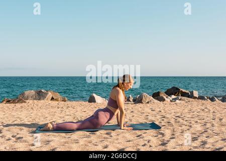 Vista laterale della donna che fa yoga a Bhujangasana e stretching corpo sulla spiaggia di sabbia in giornata di sole Foto Stock