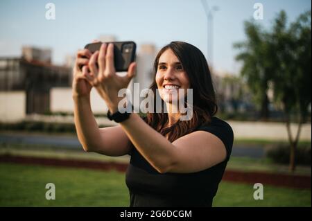 Atleta latina sorridente che prende selfie con il telefono cellulare mentre si siede sul tappetino durante l'allenamento sul prato Foto Stock