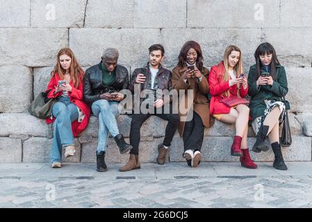 Compagnia di diversi amici seduti in città e utilizzando i telefoni cellulari mentre si agghiacciano nel fine settimana Foto Stock