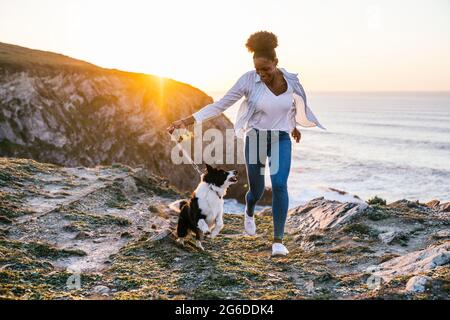 Giovane donna afroamericana che corre con il cane Border Collie mentre si passa il tempo insieme sulla spiaggia vicino al mare ondeggiante al tramonto Foto Stock