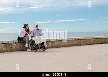 Figlia adulta e padre anziano in sedia a rotelle che si agghiacciano sul terrapieno contro il mare insieme in estate Foto Stock
