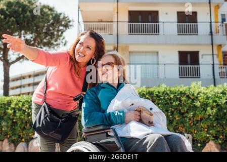 Felice donna adulta che spinge la sedia a rotelle con madre anziana mentre cammina per strada in città durante l'estate Foto Stock