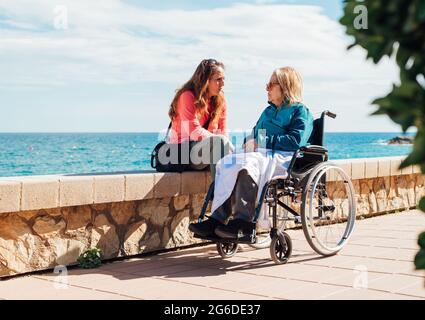 Donna adulta e madre anziana in sedia a rotelle che si aggredischettano sul terrapieno e che parlano l'una con l'altra nelle giornate di sole vicino al mare Foto Stock