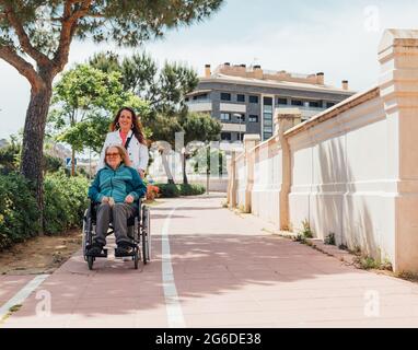Donna adulta felice guardando la macchina fotografica mentre spinge la sedia a rotelle con la madre anziana durante la passeggiata sulla strada in città in estate Foto Stock