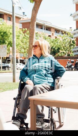 Donna anziana e pusiva seduta in sedia a rotelle sulla strada durante il giorno di sole e guardando via Foto Stock