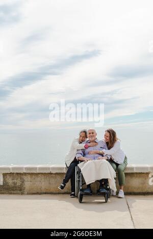Felice famiglia abbracciando padre anziano seduto in sedia a rotelle sul terrapieno vicino al mare in giornata di sole Foto Stock