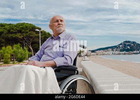 Uomo anziano in sedia a rotelle sul lungomare che guarda in contemplazione e gode di vista sul mare Foto Stock