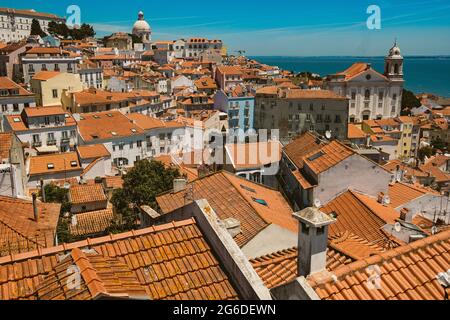 Vista panoramica dello skyline di Lisbona in una giornata estiva soleggiata con i tradizionali tetti arancioni del quartiere di Alfama con il mare visibile nel paesaggio Foto Stock