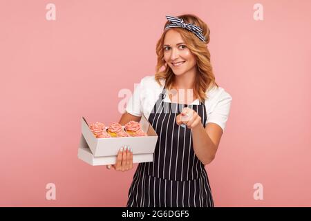 Sorridente bella donna panettiere in grembiule nero essere felice di finire ordine per la consegna, tenendo la scatola con torte e puntando il dito alla macchina fotografica. Stu. Interno Foto Stock