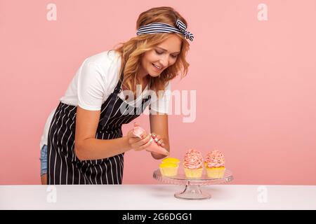 Sorridente biondo decorando muffin con crema di rose, utilizzando un cono di pasticceria, preparare l'ordine per la consegna o servire agli ospiti. studi interni Foto Stock