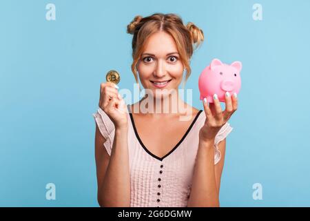 Felice femmina bionda sorridente con due ciuffi di capelli divertenti, mostrando a macchina fotografica moneta cripto oro e banca piggy, investendo in bitcoins, criptovaluta. Indo Foto Stock