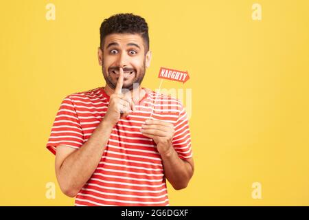 Divertente uomo bearded devious in t-shirt a righe che tiene il dito vicino alle labbra mostrando gesto shh che tiene nel bastone di mano con iscrizione naughty, sorpresa. Poll Foto Stock