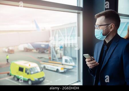Viaggiatore giovane uomo business con maschera di faccia in attesa di salire a bordo in aereo, utilizzare il telefono per il check-in aereo in aeroporto. Concetto di passeggero che viaggia con c Foto Stock