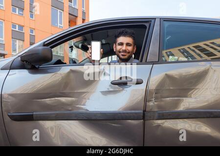 Positivo sorridente uomo bearded seduto in auto ammaccata e mostrando telefono cellulare con schermo vuoto a macchina fotografica, offre servizi di ispezione assicurativa dopo ro Foto Stock