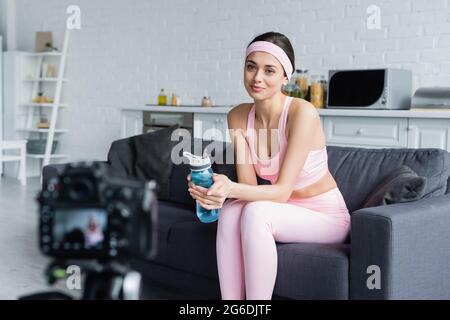 donna sorridente seduta con una bottiglia sportiva davanti a una fotocamera digitale sfocata Foto Stock