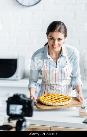 macchina fotografica digitale sfocata vicino blogger culinario felice che tiene torta gustosa Foto Stock