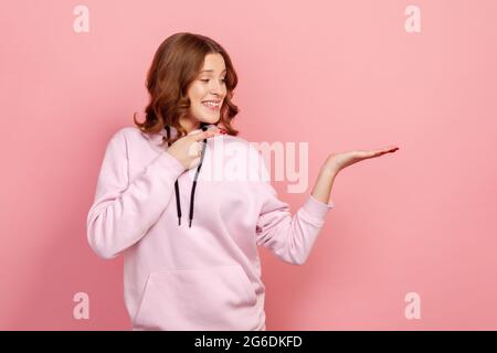 Ritratto di ragazza teen estremamente felice in hoodie che punta il dito a destra, spazio vuoto per la pubblicità, sconto. Studio al coperto isolato su rosa ba Foto Stock