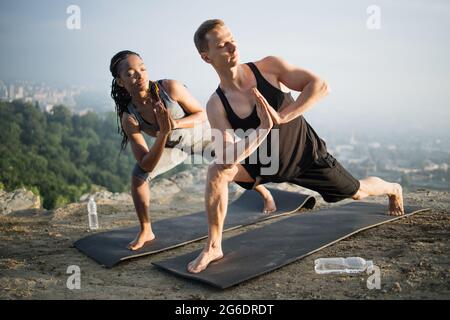 Coppia sana e forte in outfit sportivo facendo esercizi di yoga sul tappetino all'aperto. Multiculturale giovane coppia allenarsi insieme in cima al rock. Foto Stock