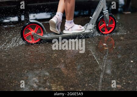 macro dettagli le gambe di una ragazza in estate scarpe cavalcare attraverso un puddle su uno scooter inviando schizzi e gocce d'acqua in diverse direzioni Foto Stock