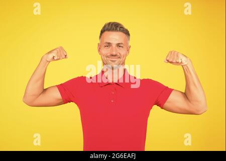 Forte sorriso atletico ragazzo in tshirt rossa flettendo braccia muscolari sfondo giallo, forza Foto Stock